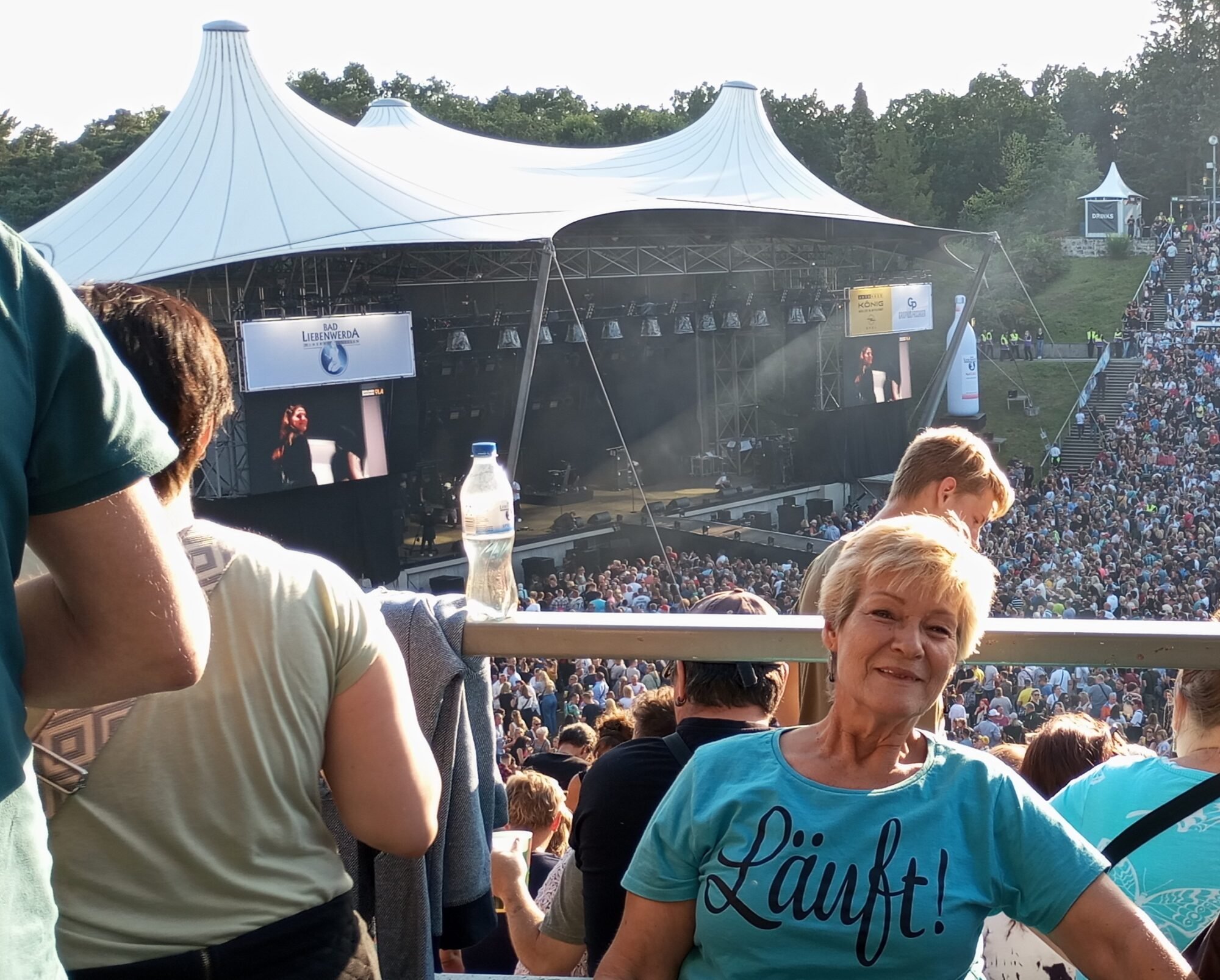 Eine Frau in einem blauen T-Shirt mit der Aufschrift "Läuft" sitzt in einem Rollstuhl am oberen Rang der Freilichtbühne der Wuhlheide auf der gerade ein Open-Air Konzert stattfindet.