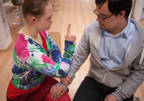 Eine junge Frau und ein junger Man mit Down-Syndrom: Er fasst sie ans Bein und sie zeigt mit dem Finger, um ein "Nein" zum Ausdruck zu bringen.