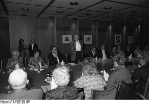 Ein historisches Schwarz-Weiß-Bild: Lieselotte Berger stehend an einem Tisch mit vielen anderen behinderten und nicht-behinderten Personen.