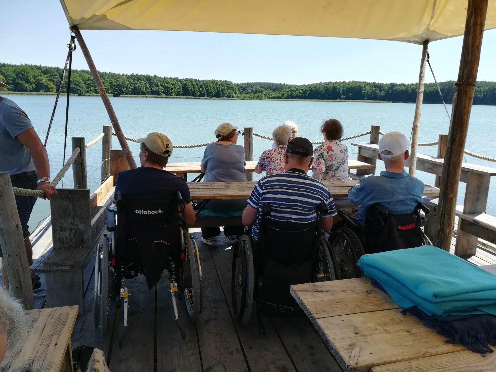 Eine Gruppe von sieben Menschen, davon drei im Rollstuhl sitzen auf einem hölzernen Floß und blicken au den See. Sie sind von hinten fotografiert, man sieht deshalb ihre Rücken. Das Wetter ist gut, das Wasser blau.