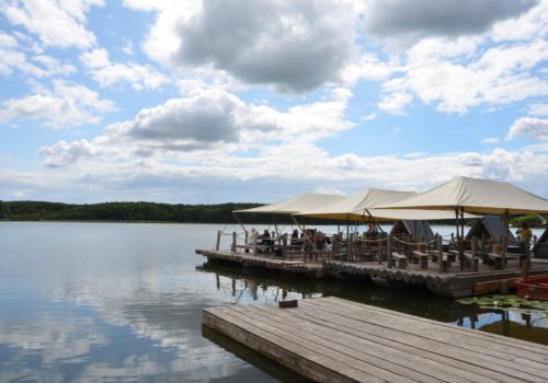 Vorne rechts ein hölzerner Steg. Dahintrer drei aneinandergebundene Flöße aus Holz mit Zeltdächern. Im Hintergrund grüner Wald, oben blauer Himmel mit vielen großen Wolken, die sich im Wasser des Sees spiegeln.