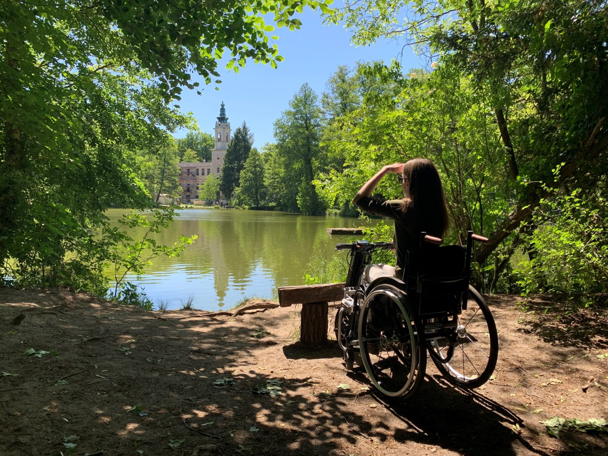 Adina sitzt mit ihrem Rollstuhl inklusive Zuggerät vor einen See im Wald und blickt in die Ferne.