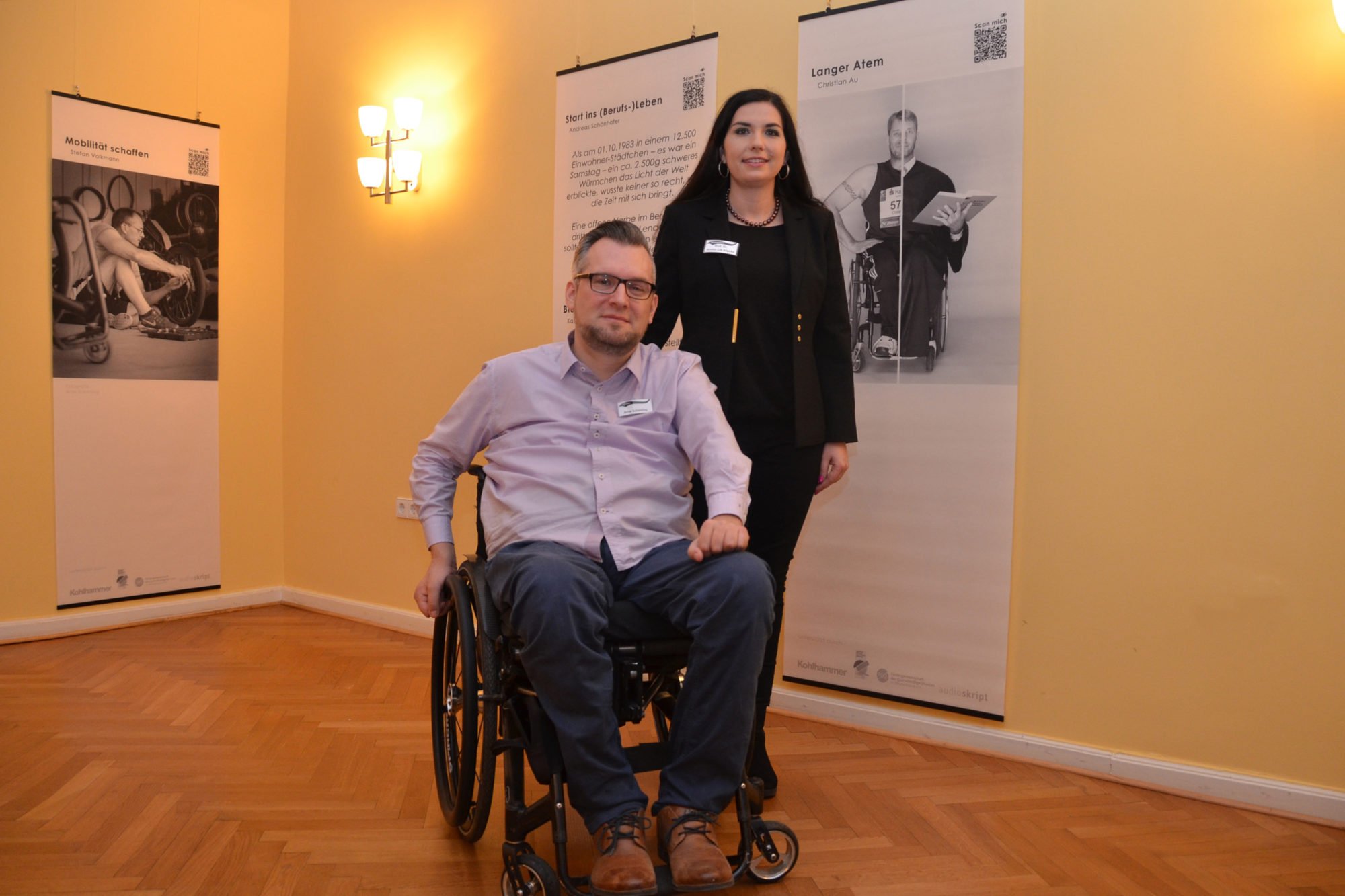 Arne Schöning vorne, im Rollstuhl, hinter ihm stehend Prof. Dr. Jessica Lilli Köpcke - im Hintergrund ist die Ausstellung zu sehen.