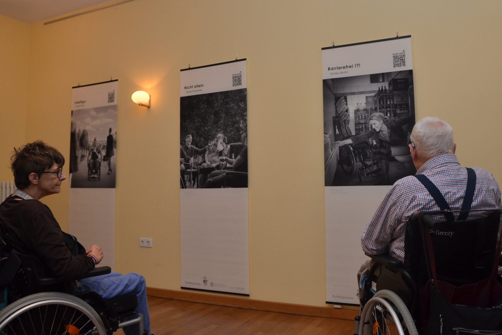 Eine Frau, links, und ein Mann, rechts, im Rollstuhl betrachten Bilder der Ausstellung.