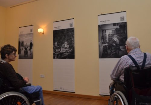 Eine Frau, links, und ein Mann, rechts, im Rollstuhl betrachten Bilder der Ausstellung.
