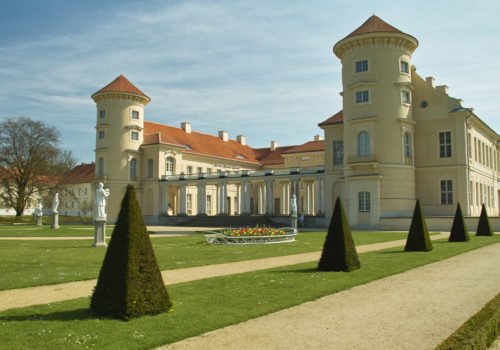 Zu sehen ist das Schloss Rheinsberg von hinten. Besonders deutlich sind die beiden Türme mit einem roten Dach und einem sandfarbenen Anstrich.