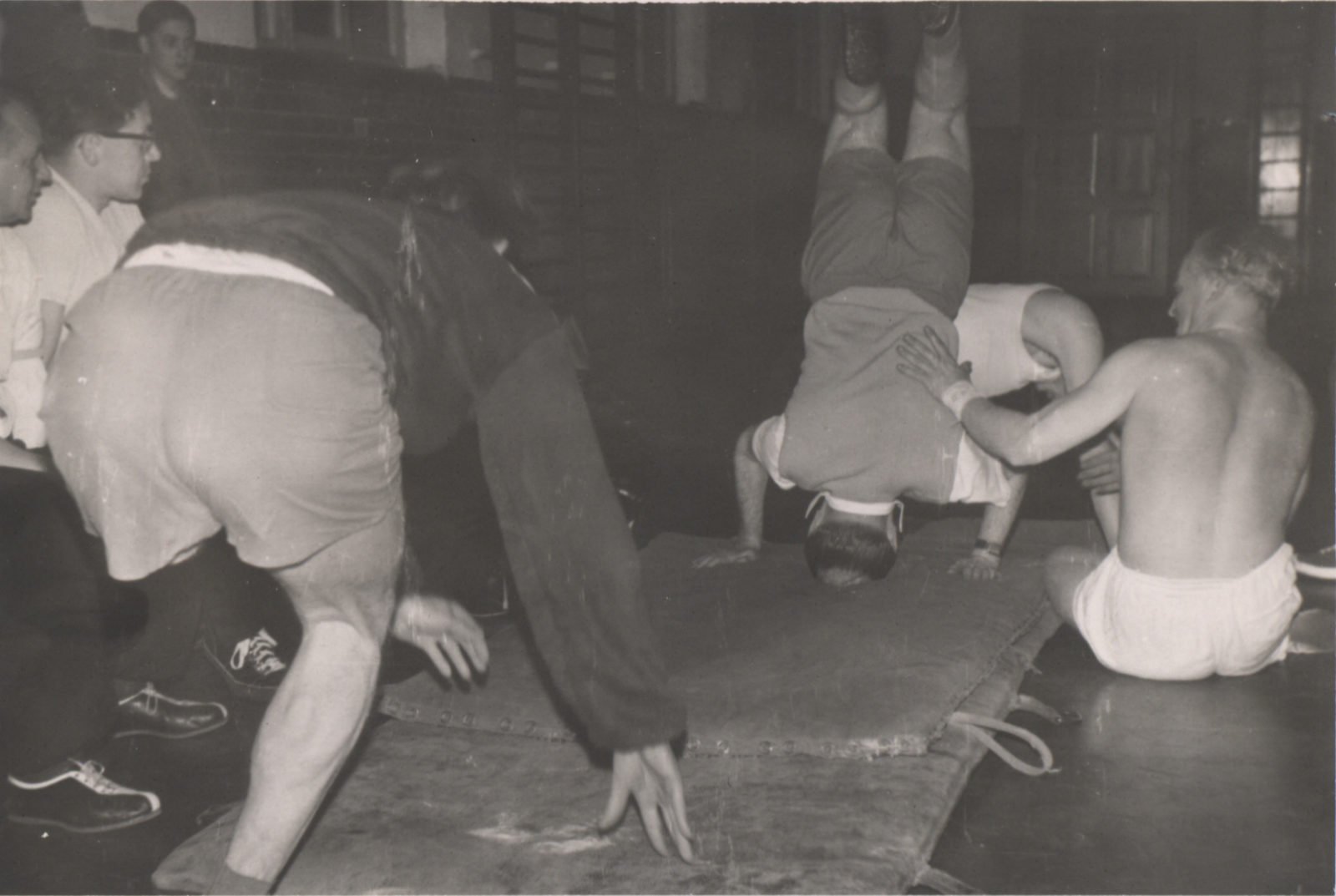Ein altes Schwarz-Weiß-Bild aus den 60er Jahren. Es zeigt mehrere Männer, die auf Bodenmatten turnen.