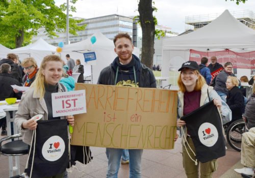 Drei junge Menschen auf dem Protesttag, in der Mitte ein junger Mann mit einem Schild auf dem "Barrierefreiheit ist ein Menschenrecht" steht, links und rechts neben ihm jeweils eine junge Dame, beide halten Beutel mit der Aufschrift I llove Vielfalt nach oben, die linke Frau außerdem ein kleineres Schild mit der Aufschrift "Barrierefreiheit ist Menschenrecht"