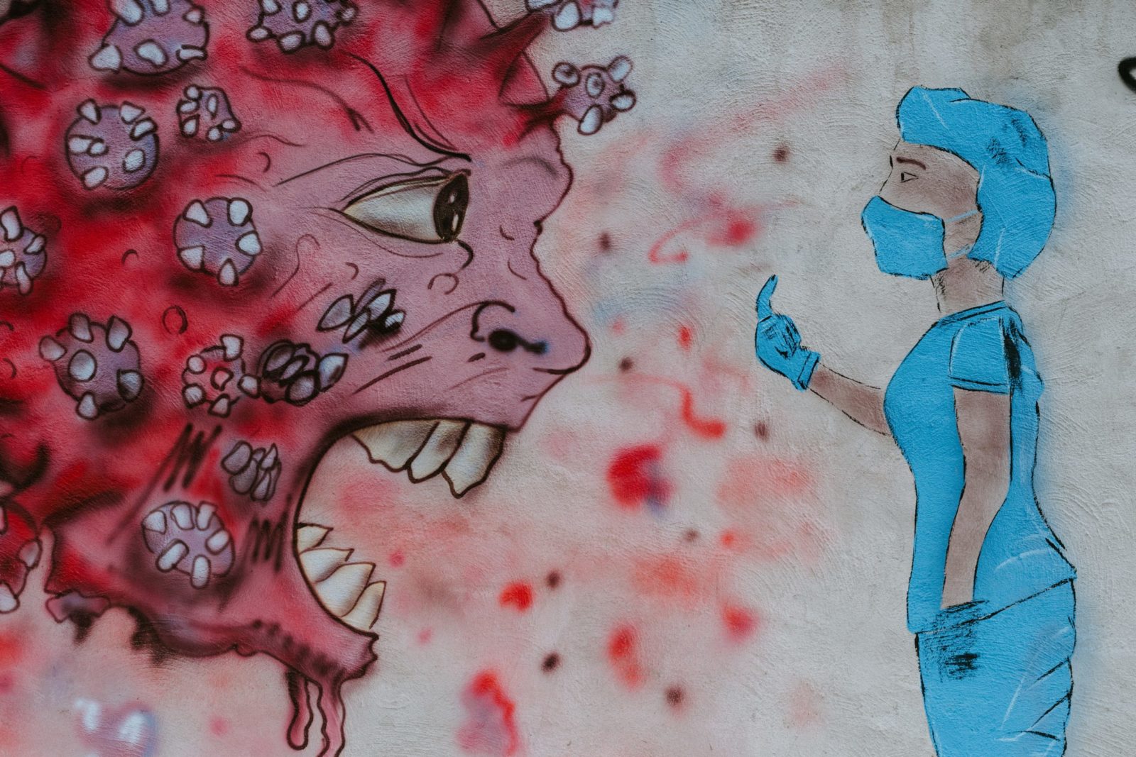Graffiti auf Hauswand: Eine in Schutzkleidung gekleidete Person zeigt einem Cartoon-Coronavirus (mit großen Augen und scharfen Zähnen) den Mittelfinger.
