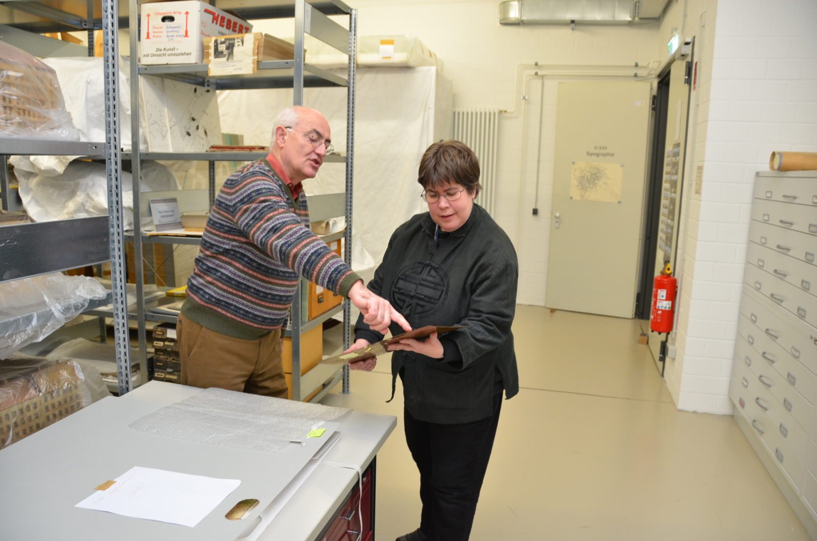 Gerd Heinemann steht links und Anke Köhler rechts im großen Depot-Raum. IM Hintergrund sind große Regale und Fächer mit Exponaten zu erkennen. Heinemann zeigt Köhler ein Exponat und deutet mit dem Finger an eine bestimmte Stelle.