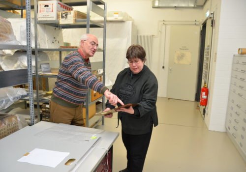 Gerd Heinemann steht links und Anke Köhler rechts im großen Depot-Raum. IM Hintergrund sind große Regale und Fächer mit Exponaten zu erkennen. Heinemann zeigt Köhler ein Exponat und deutet mit dem Finger an eine bestimmte Stelle.
