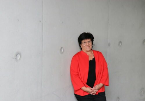 Pressefoto von Claudia Moll in rotem Blazer vor eine grauen Wand.