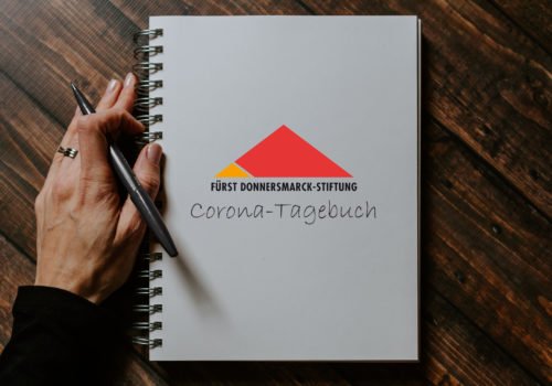 Ein Ringbuch mit FDST-Logo und der Aufschrift Corona-Tagebuch. Daneben liegt eine Hand, die einen Stift in der Hand hält.