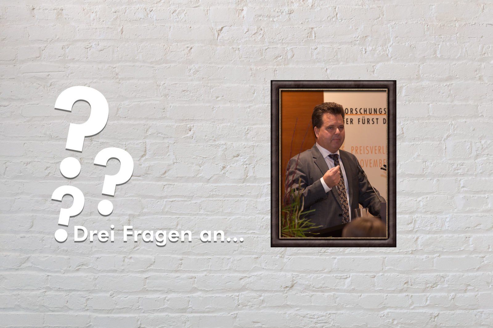 Fotomontage: Eine weiße Steinwand - darauf ein Rahmen, in den ein Bild von Jürgen Dusel montiert ist, daneben der Schriftzug Drei Fragen an... und drei Fragezeichen.