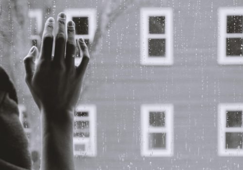 Schwarz-Weiß-Foto: Vorne eine Hand von hinten fotografiert, die an eine Fensterscheibe fasst. Im Hintergrund eine Häuserfassade mit vielen Fenstern.