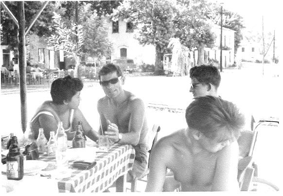 Scan einer Schwarzweißfotografie: drei Männer, alle oberkörperfrei und einer davon im Rollstuhl sitzend, und eine Frau sitzen gemeinsam an einem Tisch.