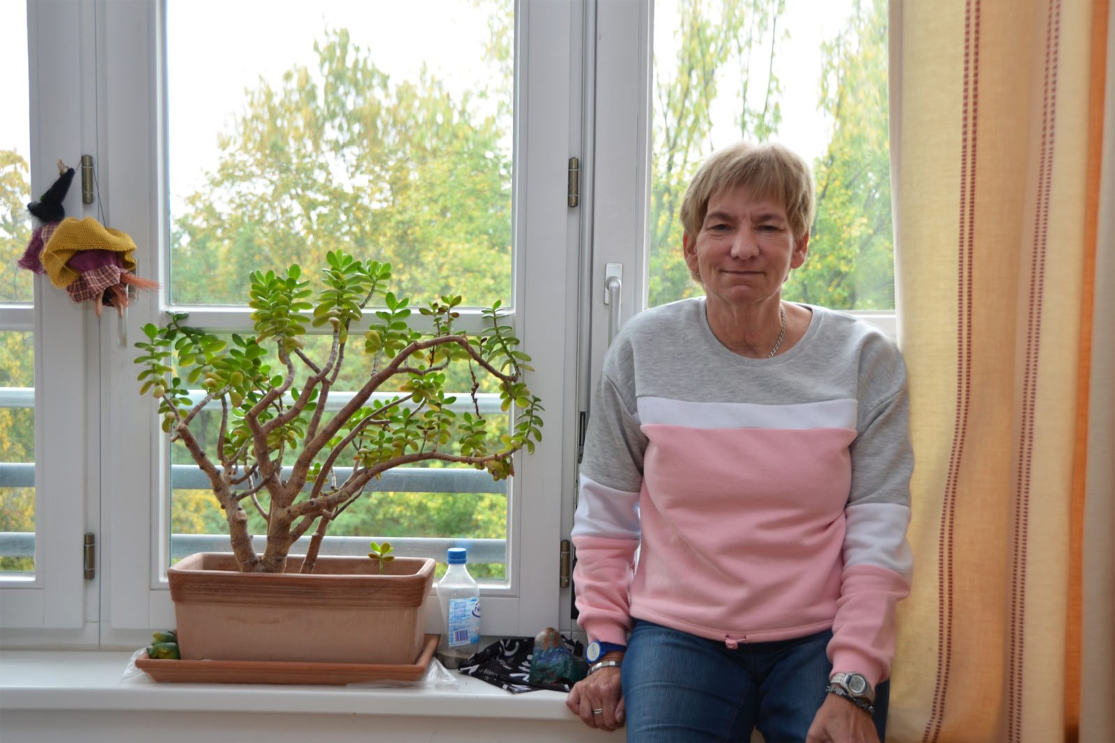Barbara Hagedorn angelehnt an ihrer Fensterbank. Neben ihr eine Pflanze. Im Fenster hängt eine Hexe aus Stoff.