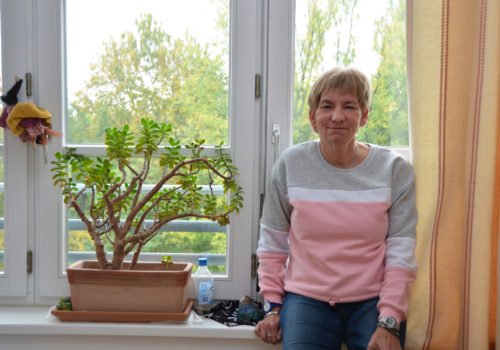 Barbara Hagedorn angelehnt an ihrer Fensterbank. Neben ihr eine Pflanze. Im Fenster hängt eine Hexe aus Stoff.