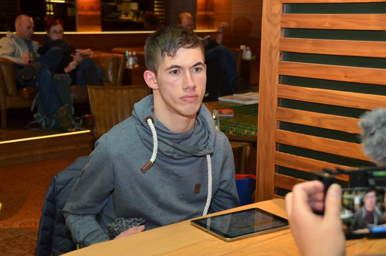 Jasper Dombrowski sitzt an einem Tisch in einem Café er wird gefilmt, das Display der Kamera ist im rechten unteren Bildrand zu erkennen, Jasper blickt direkt in die Kamera.
