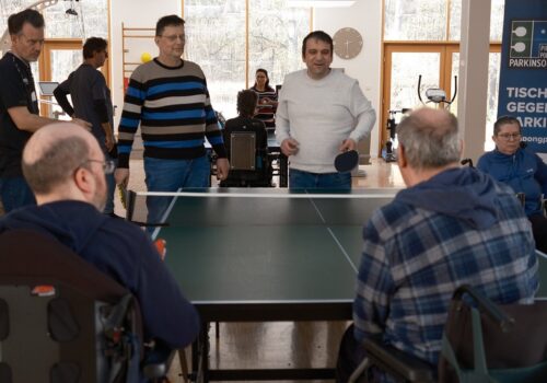Um eine Tischtennisplatte befinden sich5 Menschen. Drei von Ihnen stehen an einem Ende und zwei weitere Sitzen im Rollstuhl auf der Gegenüberliegenden Seite. Das Bild zeigt inklusiven Sport.