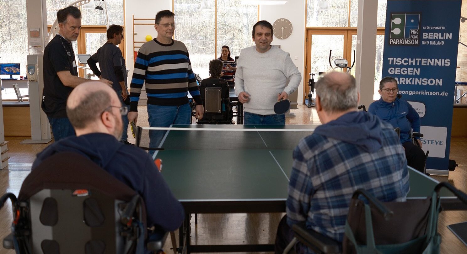Um eine Tischtennisplatte befinden sich5 Menschen. Drei von Ihnen stehen an einem Ende und zwei weitere Sitzen im Rollstuhl auf der Gegenüberliegenden Seite. Das Bild zeigt inklusiven Sport.