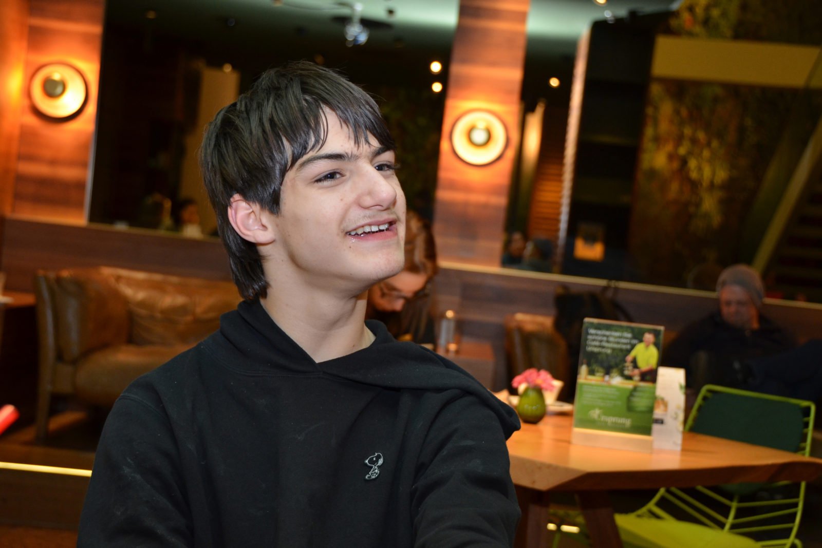 Linus Bade in einem Café - er lächelt und blickt leicht an der Kamera vorbei. Im Hintergrund sind Leute an Tischen zu erkennen, aber verschwommen.