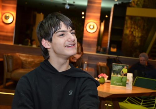 Linus Bade in einem Café - er lächelt und blickt leicht an der Kamera vorbei. Im Hintergrund sind Leute an Tischen zu erkennen, aber verschwommen.