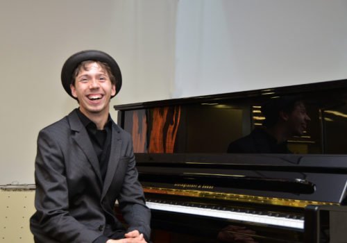 Junger Mann mit Hut sitzt vor einem Klavier und lacht in die Kamera