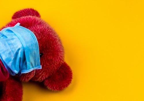 Ein roter Teddybär mit blauer OP-Maske auf gelben Hintergrund.