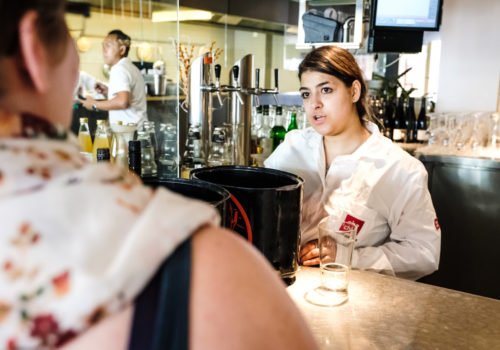Eine junge Frau steht in einem Restaurant hinter der Bar und bedient einen Gast.