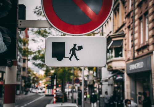 Ein Straßenschild für Halteverbot, darunter ein weißes Schild mit dem Piktogramm eines Paketboten, das symbolisiert, dass Paket- und Briefboten ausgenommen sind.