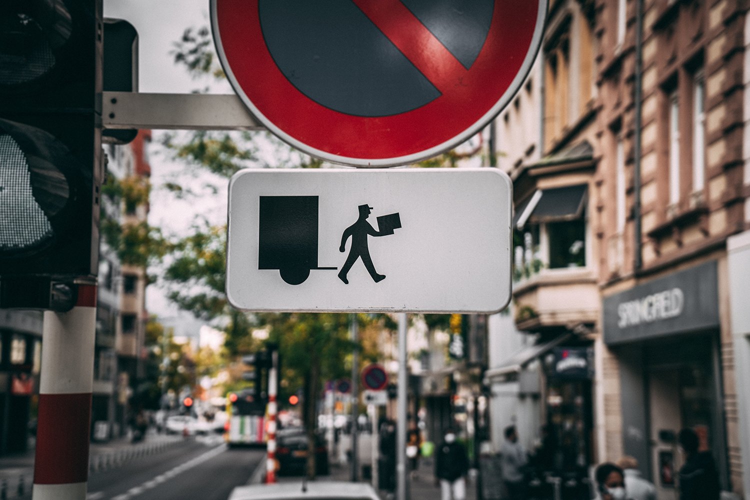 Ein Straßenschild für Halteverbot, darunter ein weißes Schild mit dem Piktogramm eines Paketboten, das symbolisiert, dass Paket- und Briefboten ausgenommen sind.