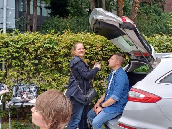 Ein Mann sitzt unter der geöffneten Kofferraum eines silbernen Autos und wird von einer Frau geschminkt
