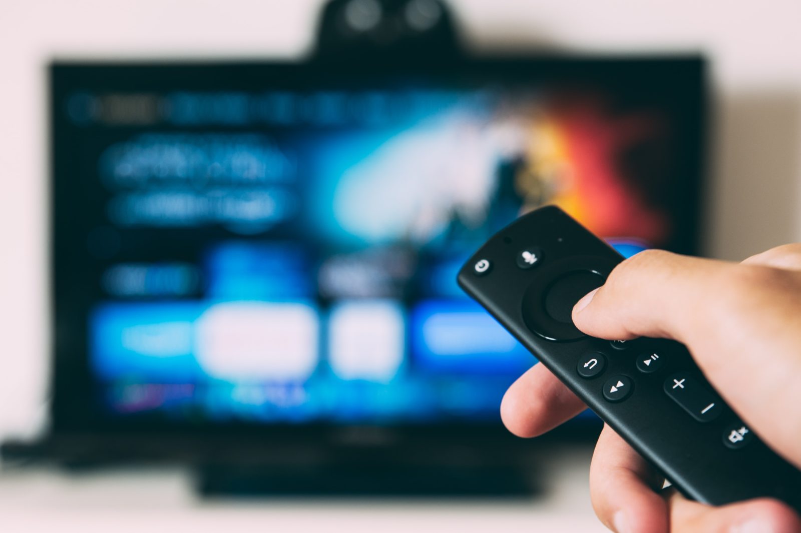 Eine Fernbedienung ist auf einen Fernseher gerichtet, der im Hintergrund nur verschwommen zu erkennen ist. Wahrnehmbar ist allerdings, dass ein Streaming-Dienst zu sehen ist.