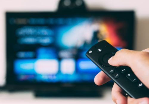 Eine Fernbedienung ist auf einen Fernseher gerichtet, der im Hintergrund nur verschwommen zu erkennen ist. Wahrnehmbar ist allerdings, dass ein Streaming-Dienst zu sehen ist.